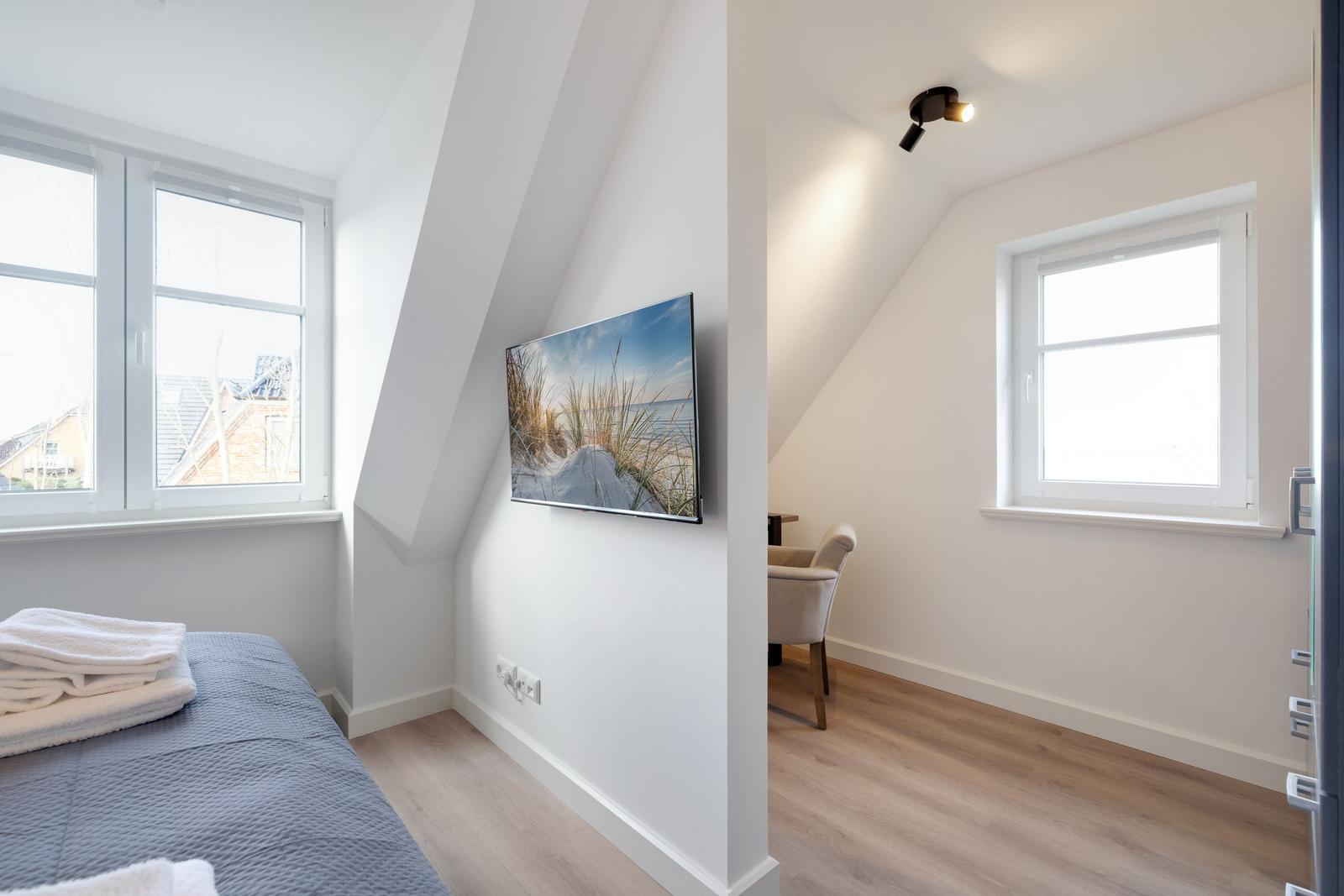 Schlafzimmer mit Dachschräge, Fenster und TV. Nebenraum mit Sessel und Fenster.