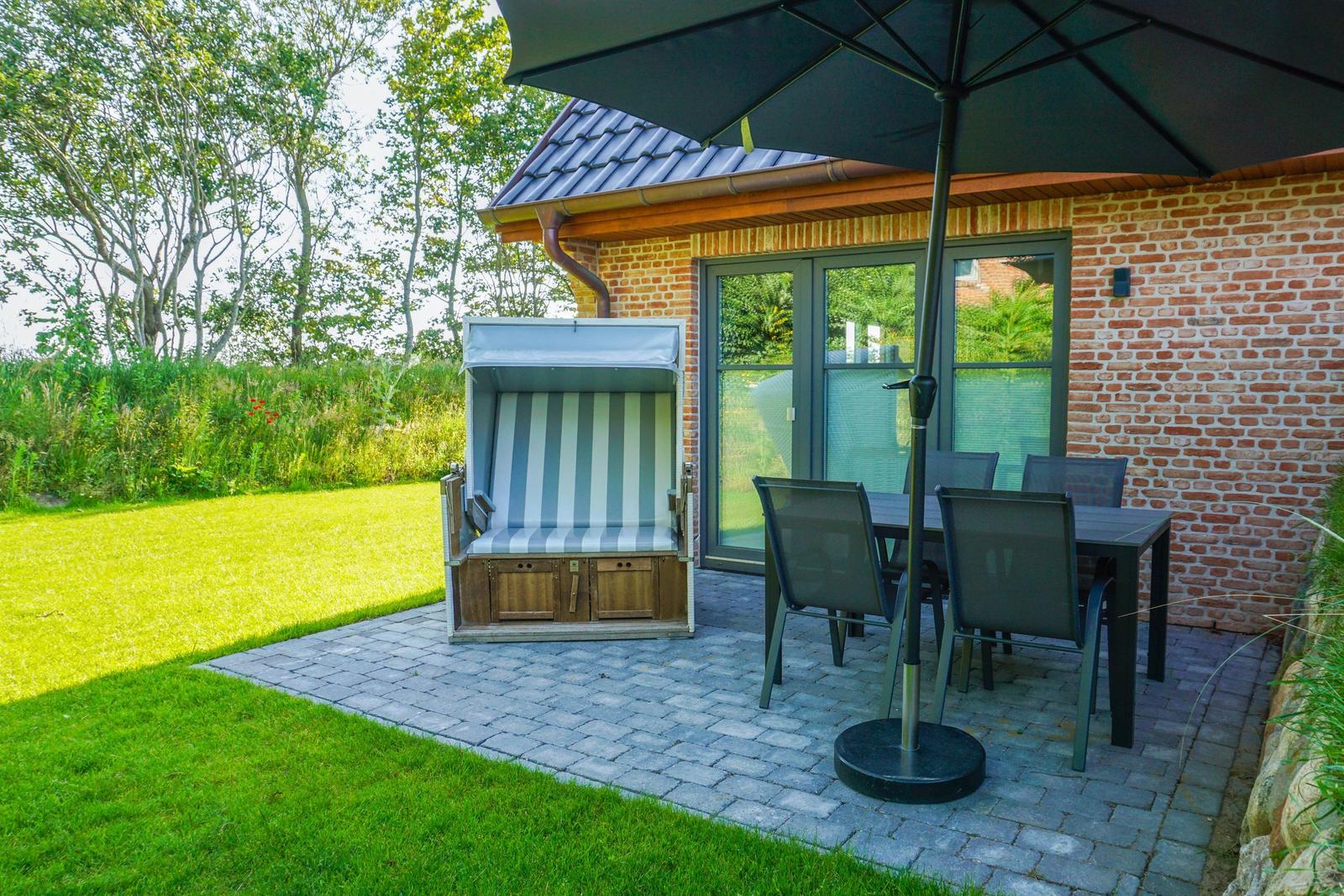 Terrasse mit Gartenliege, Tisch und Stühlen unter Sonnenschirm. Hintergrund: Grasfläche und Bäume.