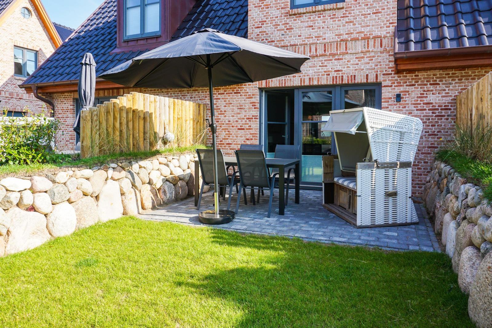 Terrasse mit Tisch, Stühlen und Sonnenschirm neben Haus. Weiße Strandkorb neben Steinmauer.