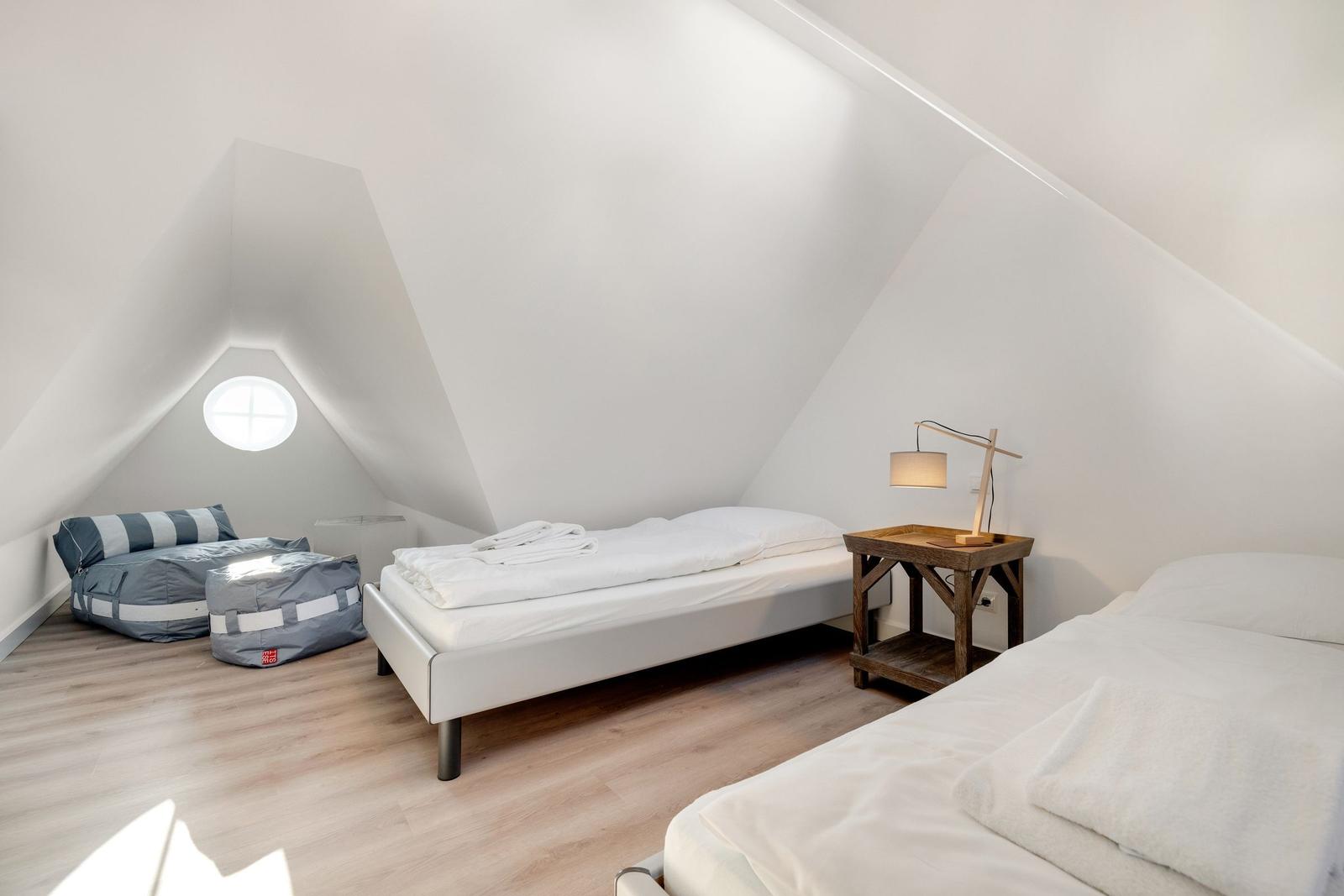 Zimmer mit zwei Betten, Holzboden und runder Fensteröffnung unter Dachschrägen.