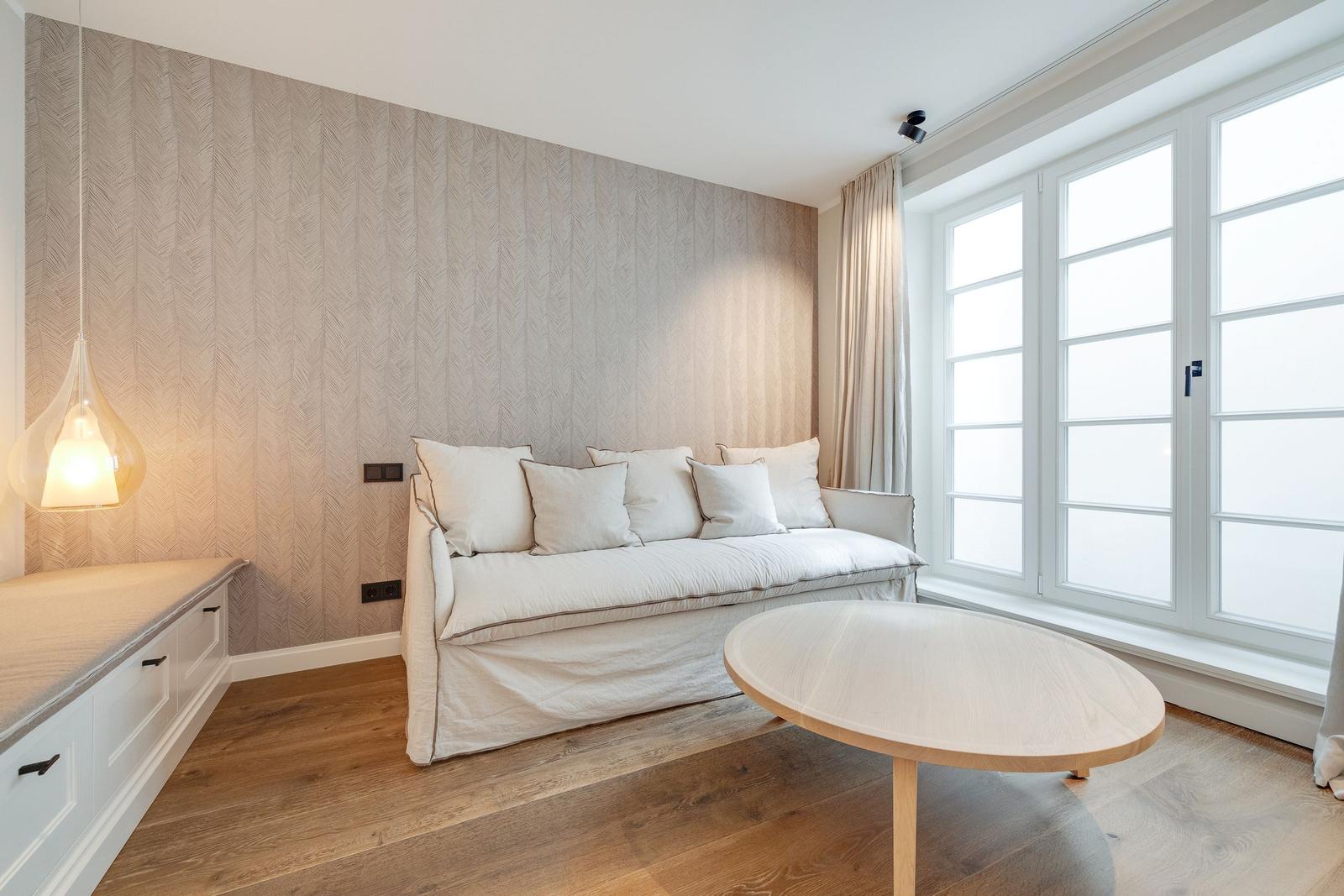 A room with sofa, round table, and large window. Patterned wall, wooden floor, and pendant lamp.