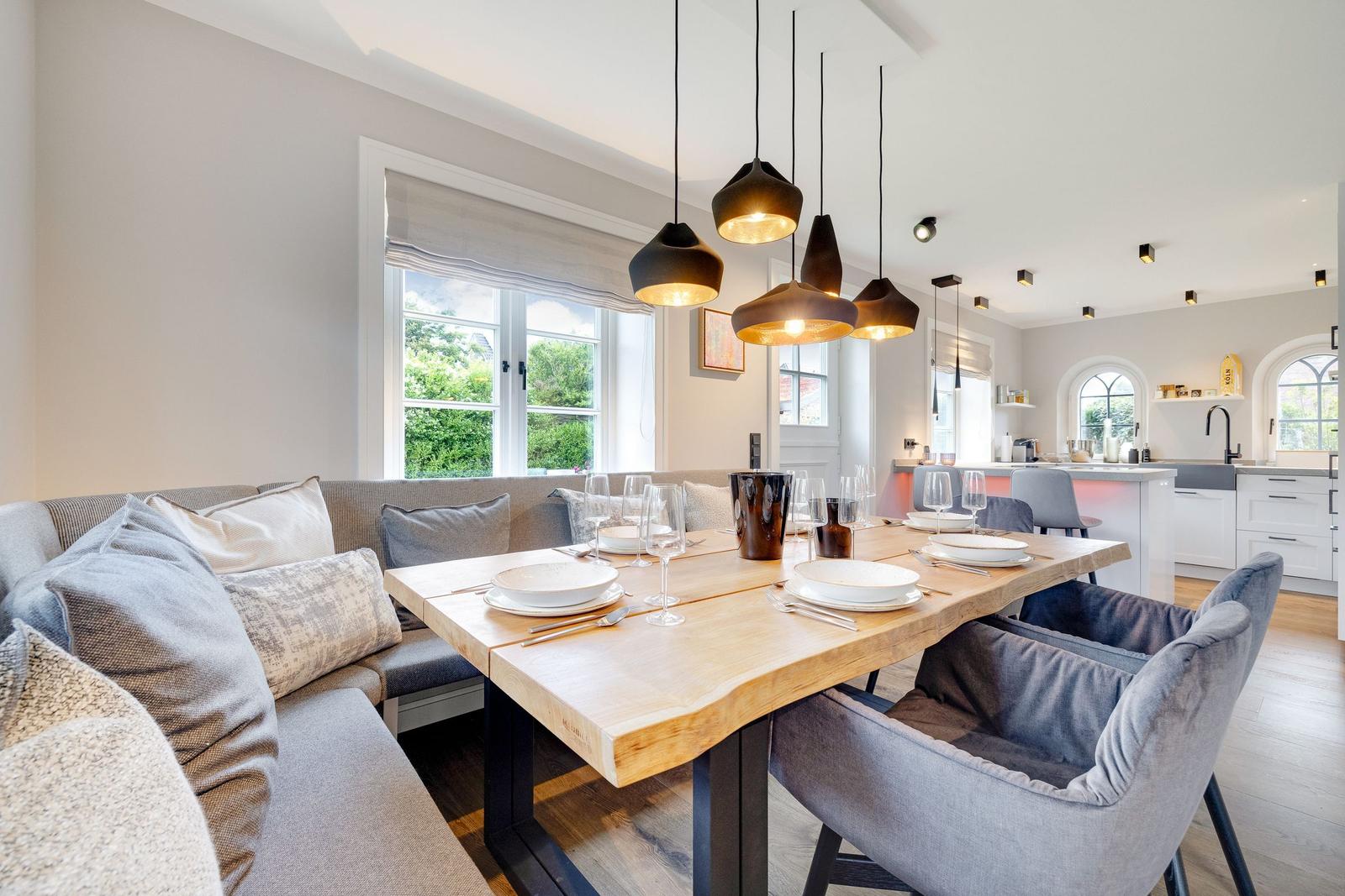 Dining area with wooden table, bench, and pendant lights. Kitchen in background.