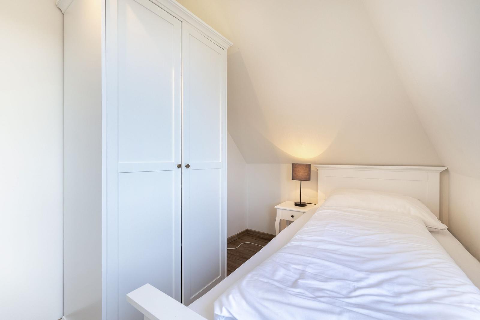Schlafzimmer mit weißem Bett, Nachttisch und Kleiderschrank unter schrägem Dach.