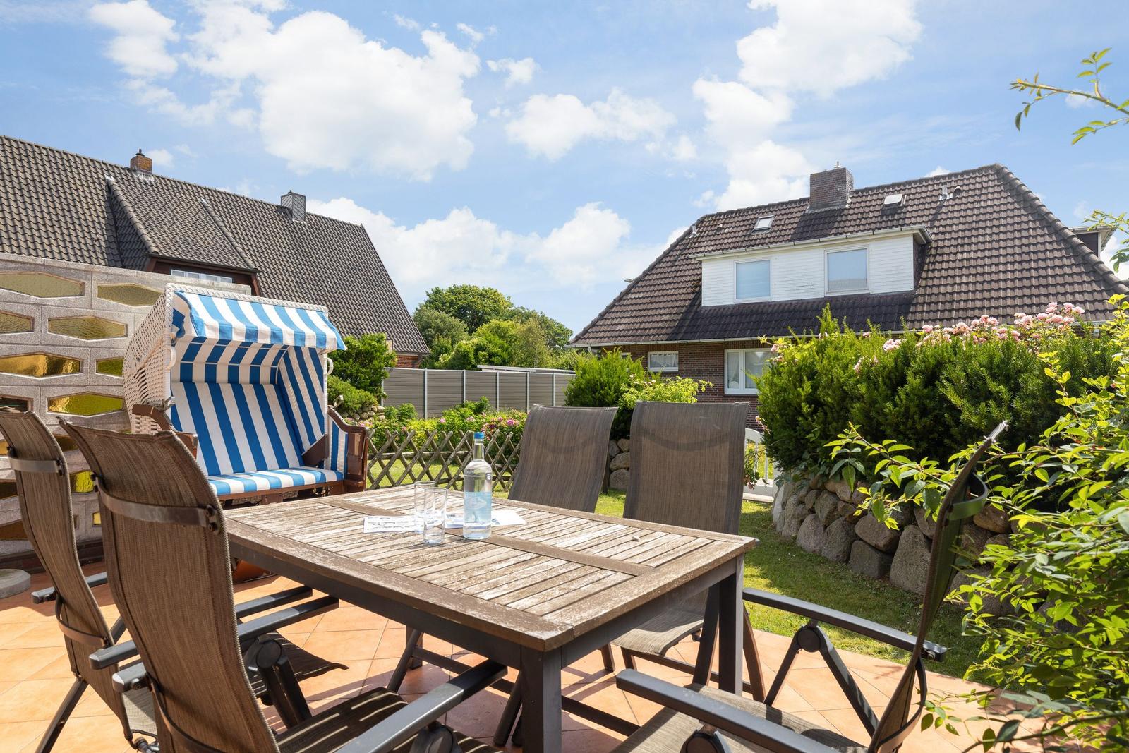 Terrasse mit Tisch, Stühlen und Strandkorb. Hintergrund: Häuser und Grünanlagen.