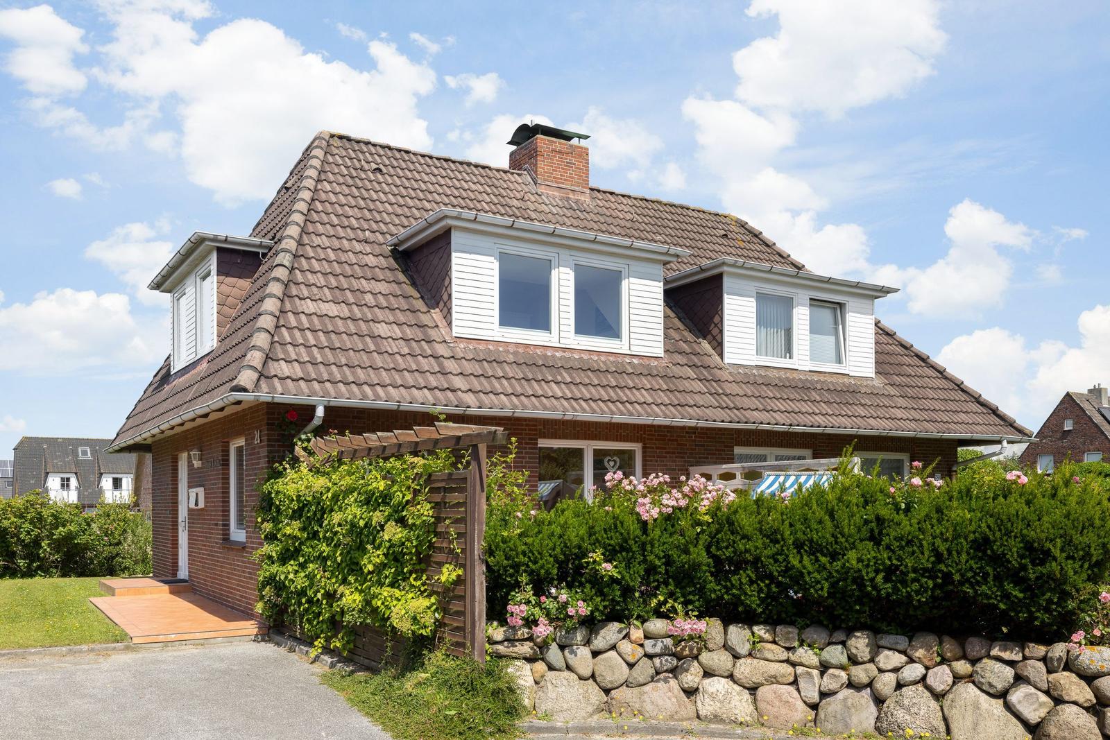 Haus mit Dachgauben, Steinmauer und Blumenbeete im Vorgarten.