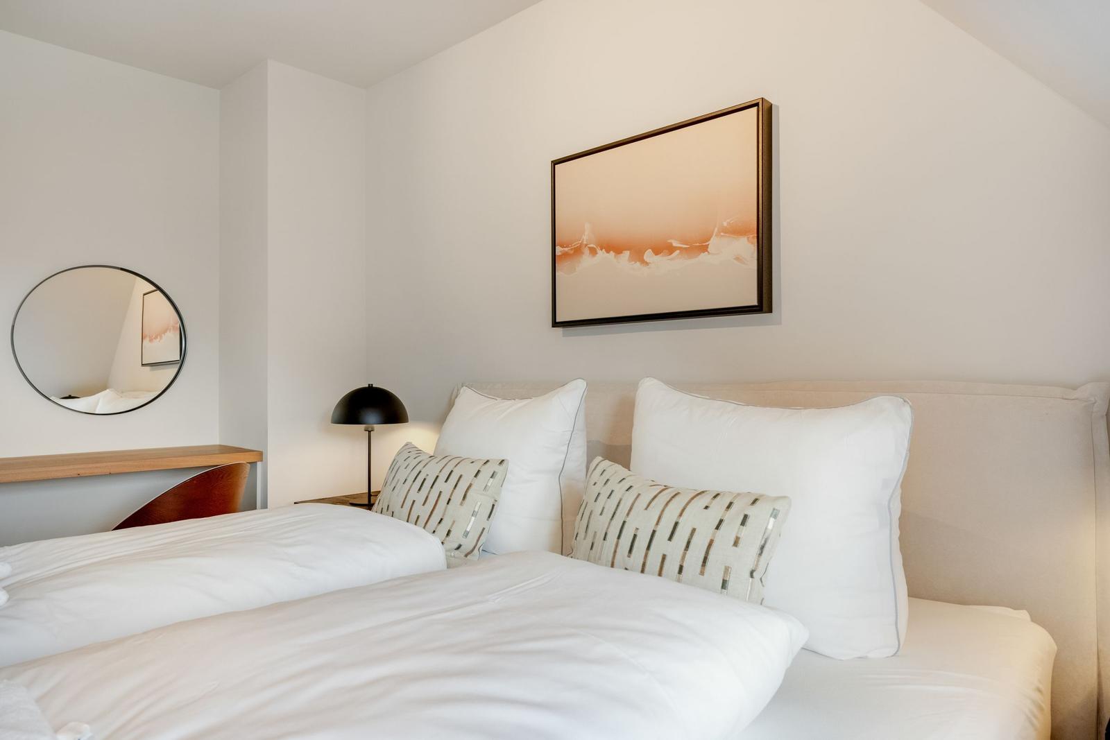 Schlafzimmer mit Bett, Kissen, Wandbild und runder Spiegel an der Wand.