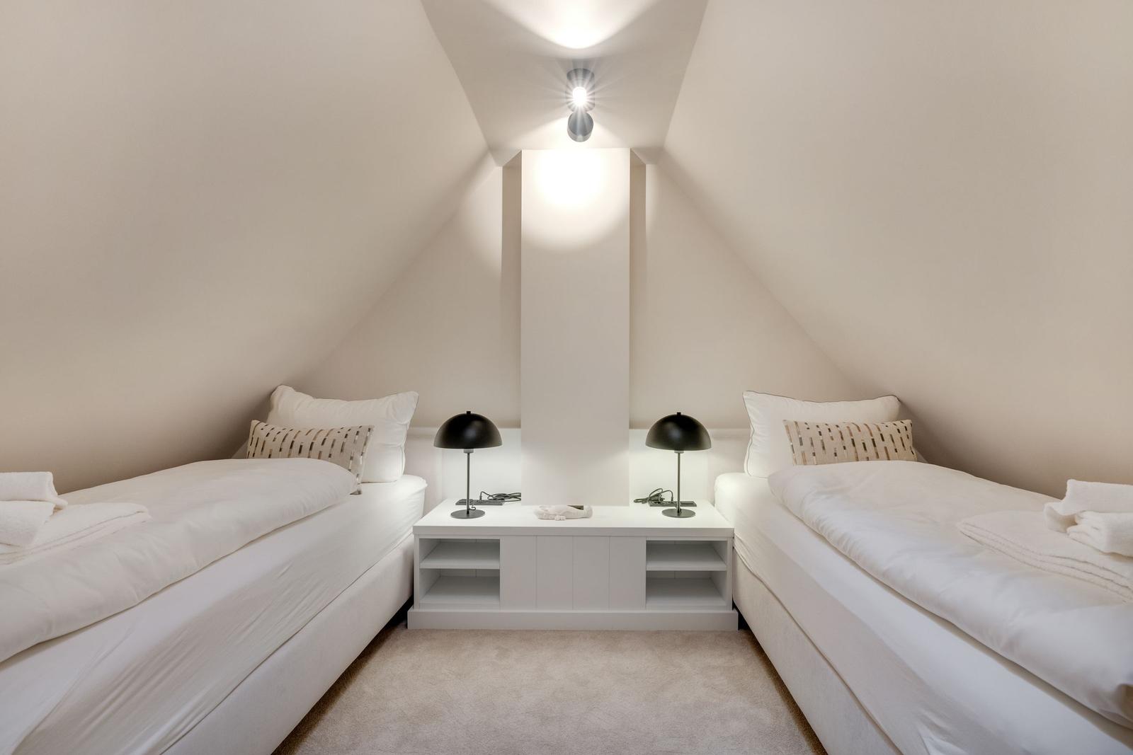 Zwei Betten mit weißen Laken und Kissen, schwarze Nachttische mit Lampen, schräges Dach und weißer Wand.