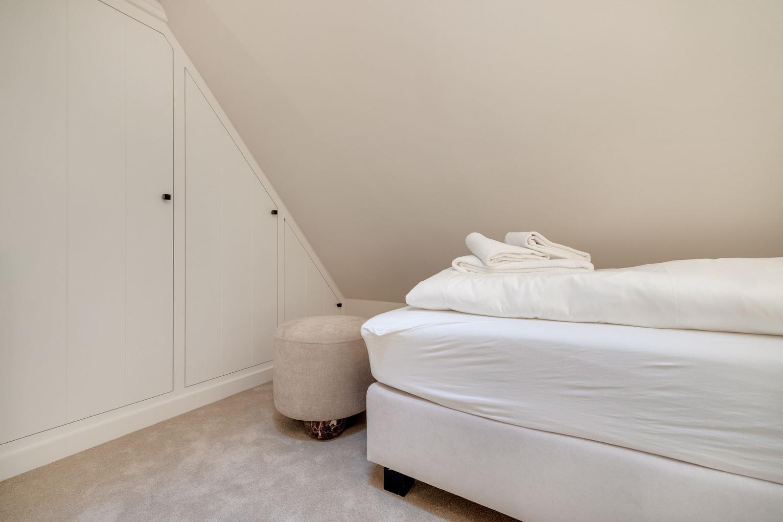 Schlafzimmer mit weißem Bett, Kleider schrank und Sitzhocker unter schräger Decke.