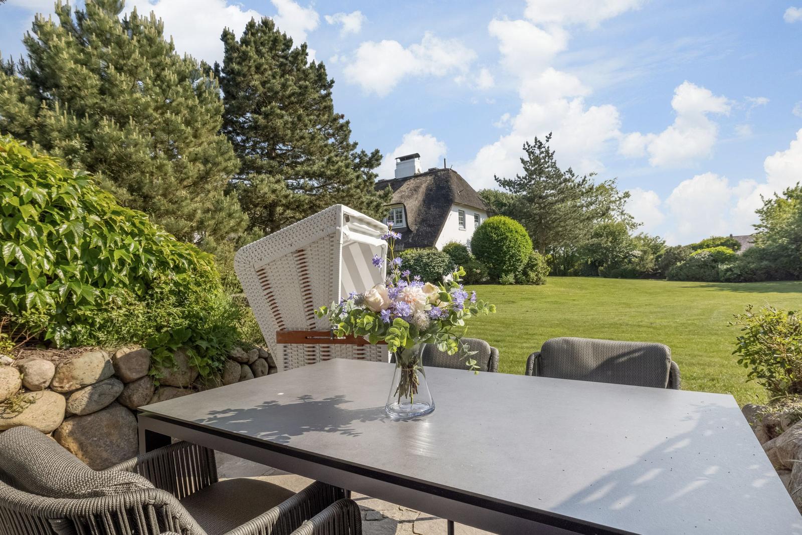 Außenbereich mit Tisch, Stühlen und Blumenvase. Blick auf grünen Rasen und Haus im Hintergrund.