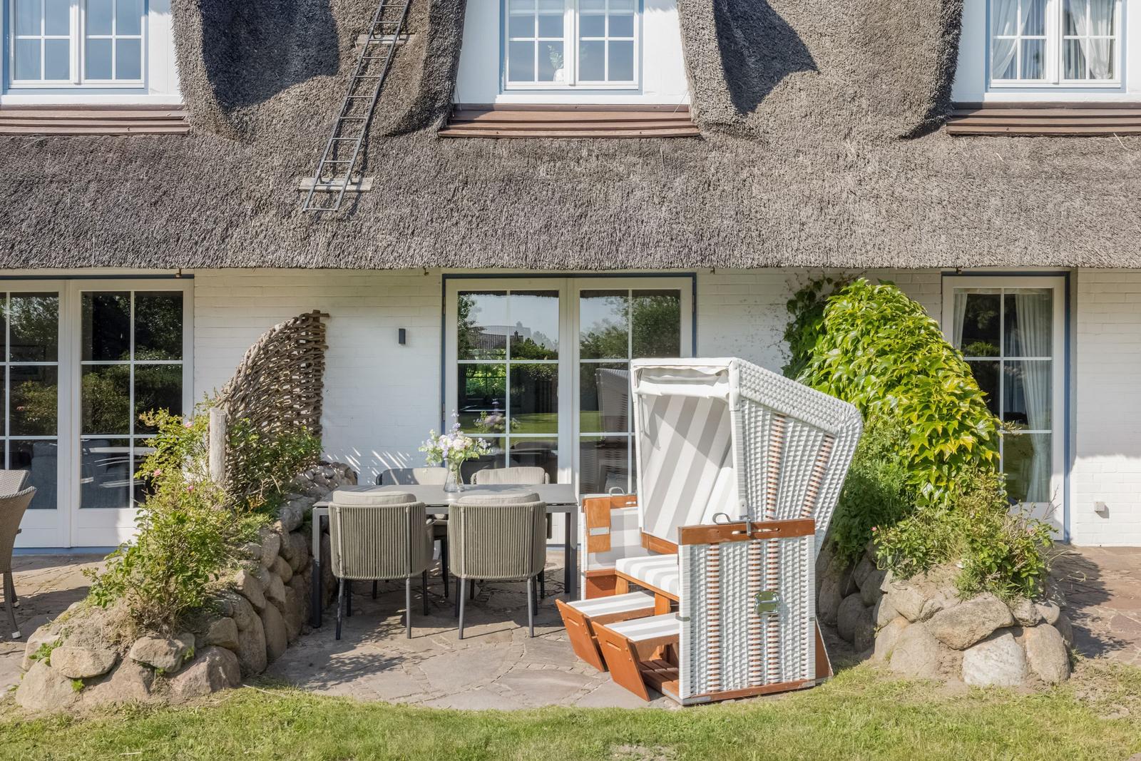 Terrasse mit Tisch, Stühlen und Strandkorb vor einem strohgedeckten Haus.