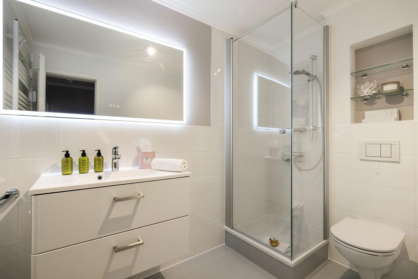 Badezimmer mit Dusche, Waschbecken und Toilette. Mit beleuchtetem Spiegel und grünen Flaschen.