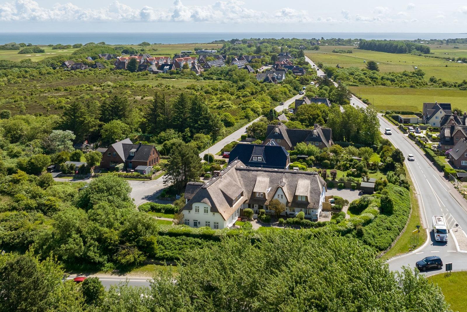 Haus mit Dachrinne, umgeben von Grünflächen und Straße. Blick auf Meer und Dorf.