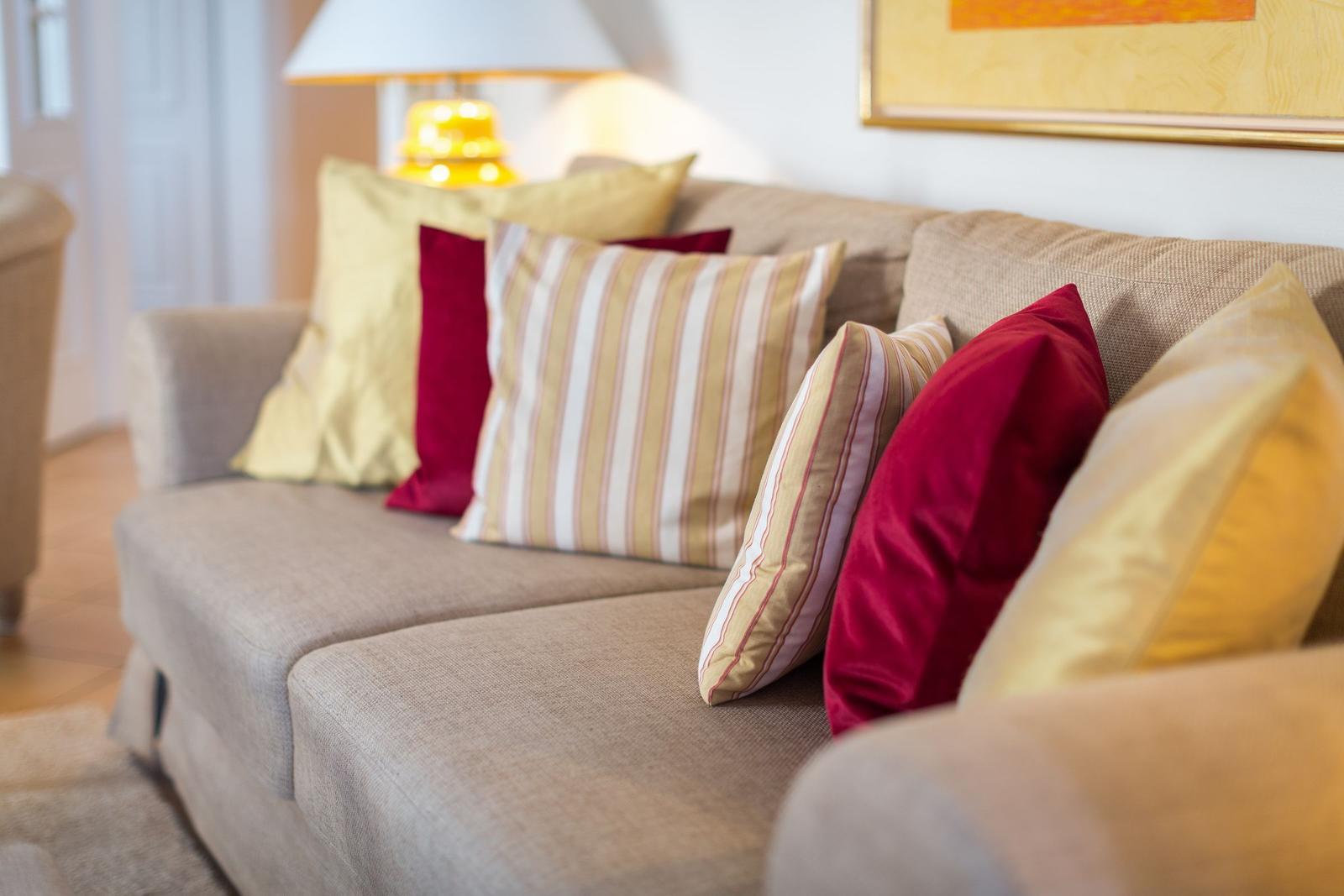 Ein Sofa mit mehreren Kissen in Rot und Gelb. Eine Lampe und ein Bild sind im Hintergrund sichtbar.
