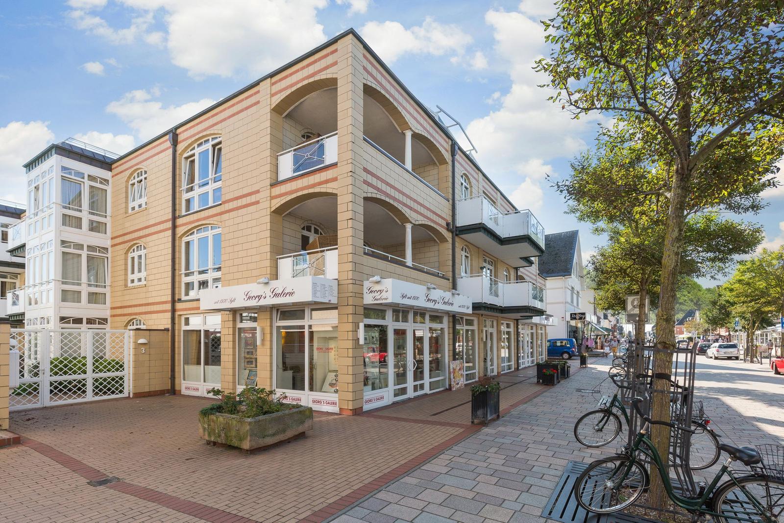 Gebäude mit Laden und Balkonen an einer Straße mit Bäumen und Fahrrädern.