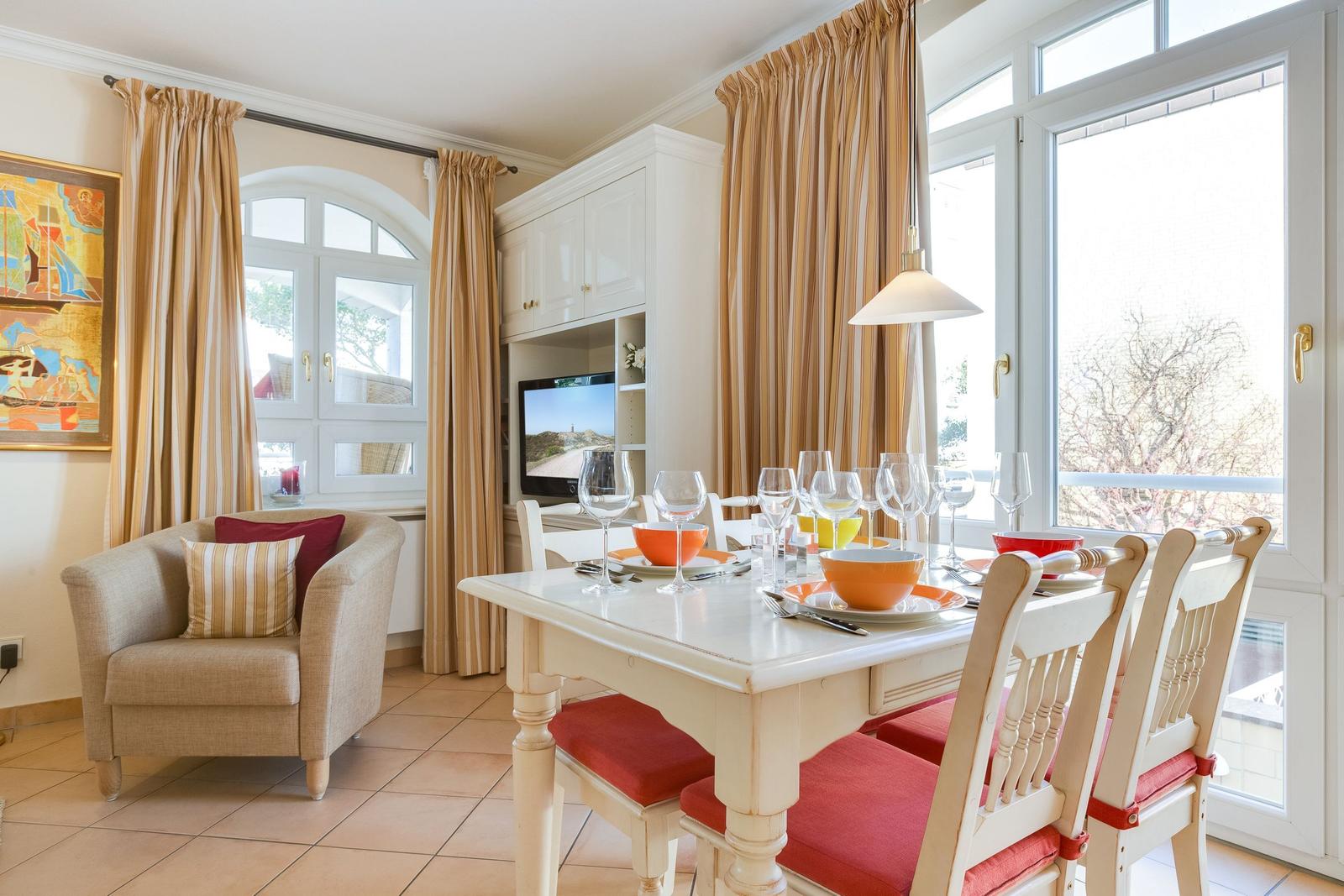 Esszimmer mit Tisch, Stühlen und Fenstern. Fernseher und Sessel im Hintergrund.