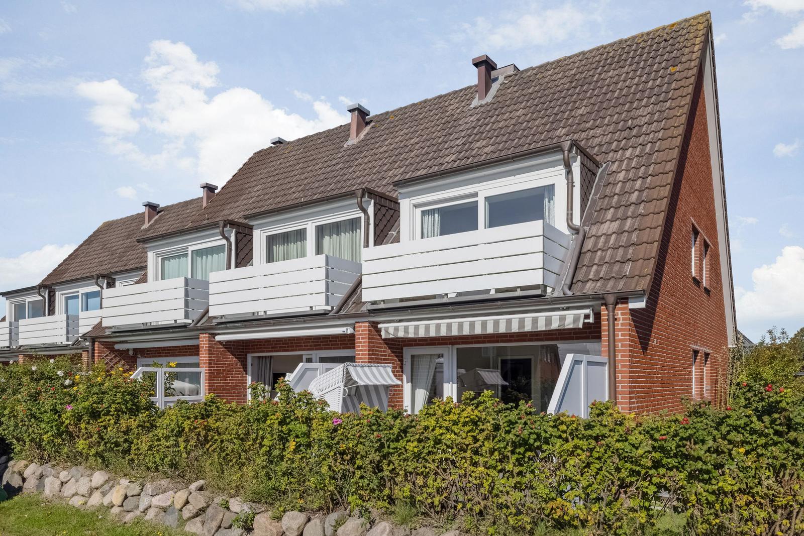 Red brick house with white balconies and garden terrace under blue sky.