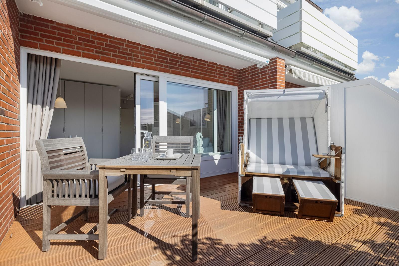 Deck with wooden table, chairs, and beach chair. Background: living area with window.