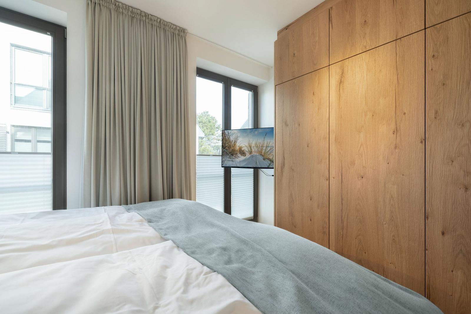 Bedroom with bed, wooden wardrobe, and TV. Windows with blinds and curtains.