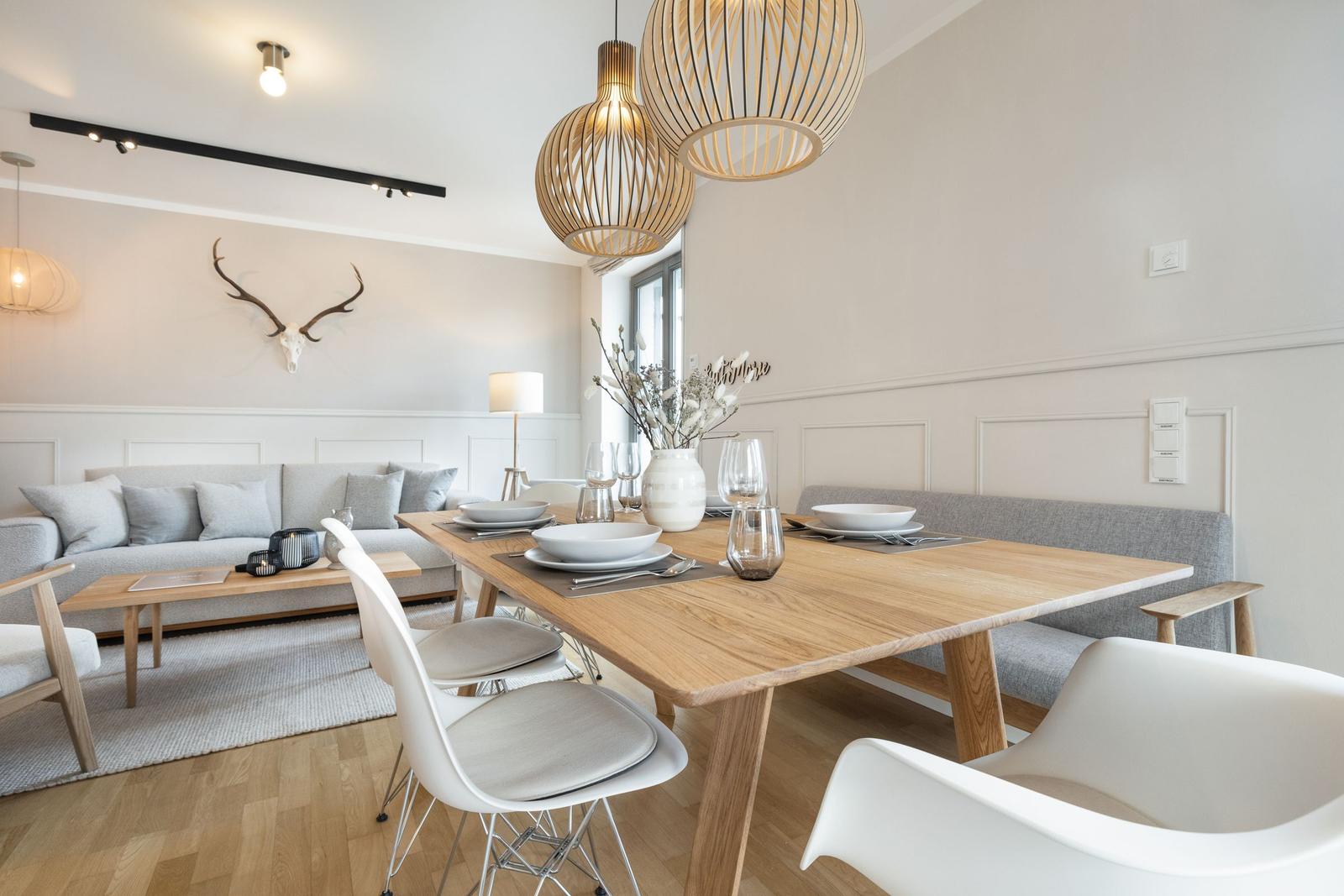 Dining area with wooden table and white chairs, next to living area with sofa and deer head on wall.
