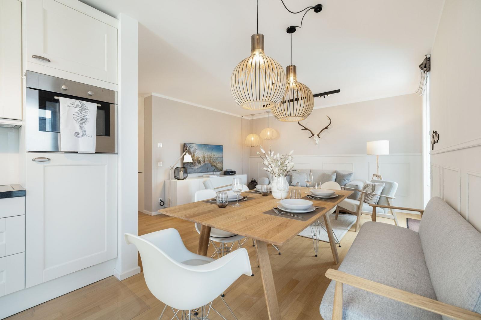 Dining room with wooden table, white chairs, and modern pendant lights. Kitchen area with built-in oven.