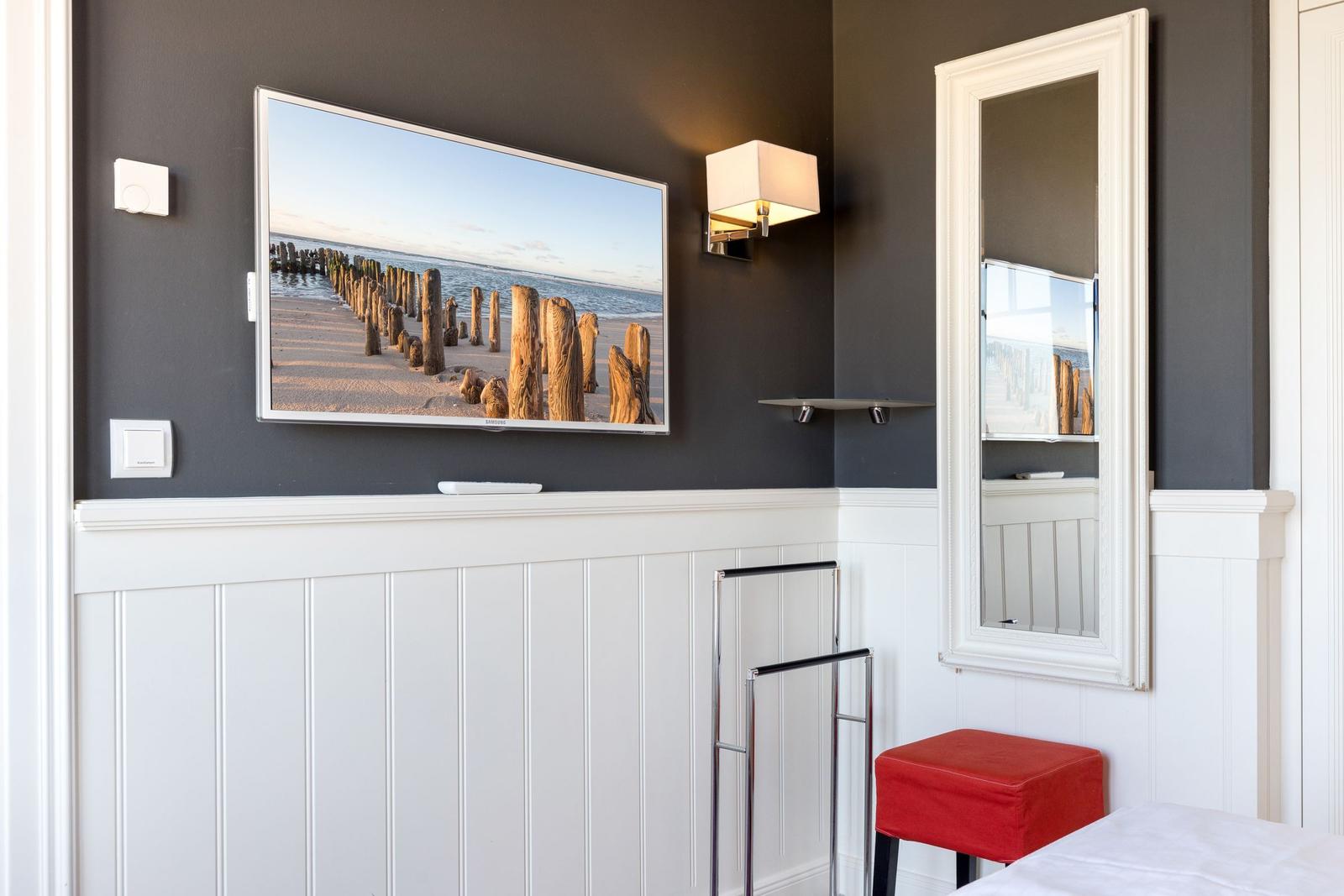 TV mounted on wall, mirror and red stool in room.