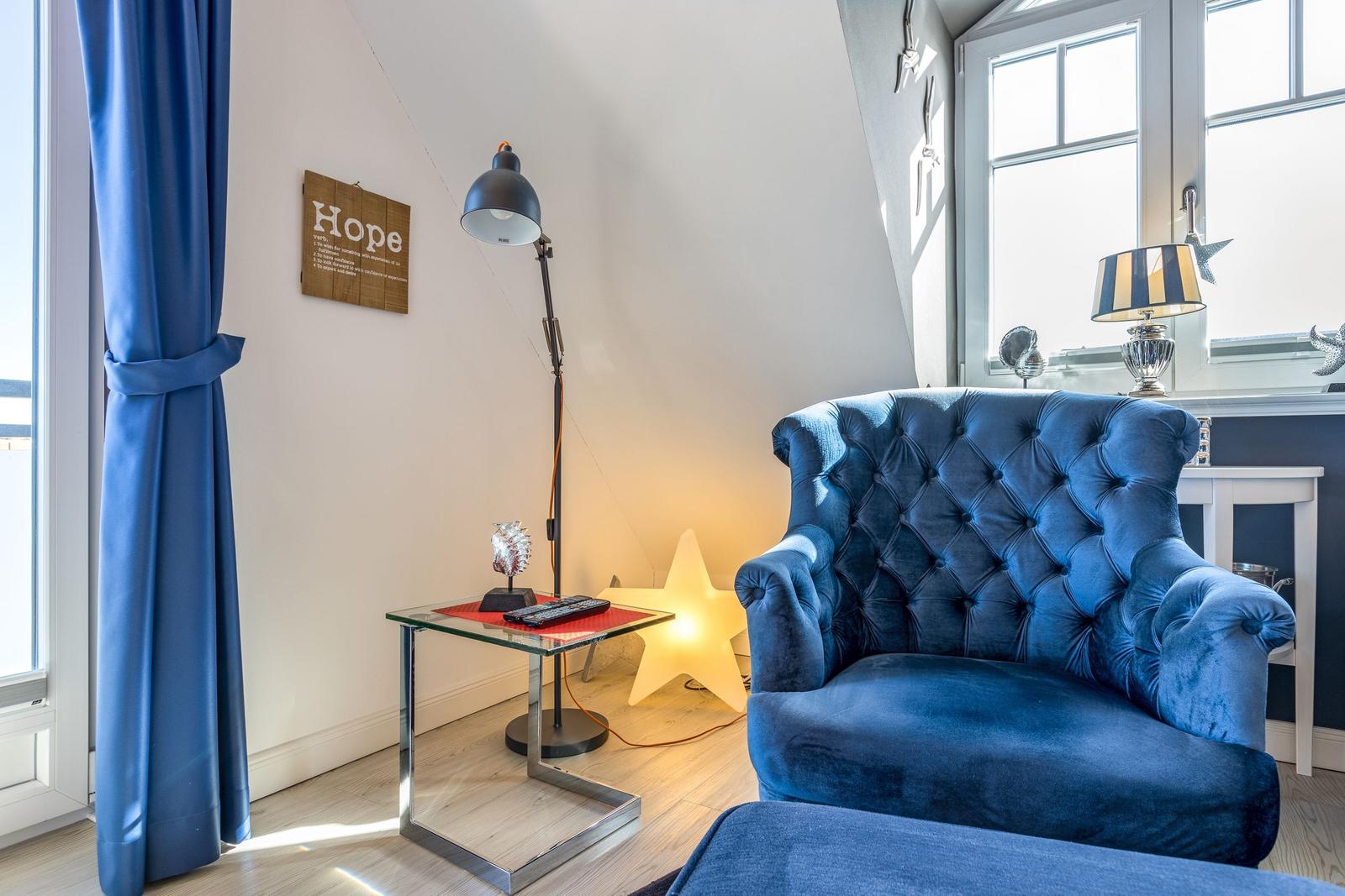 A room with blue armchair, windows and table. Wall art and lamps are visible.