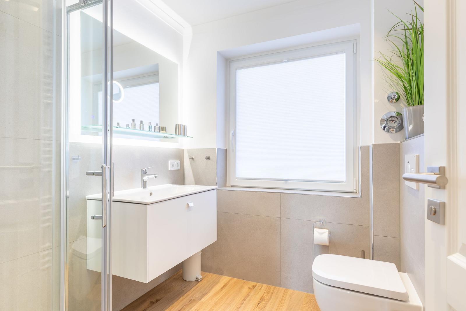 Bathroom with shower, sink and toilet. Wooden floor and window with blinds.