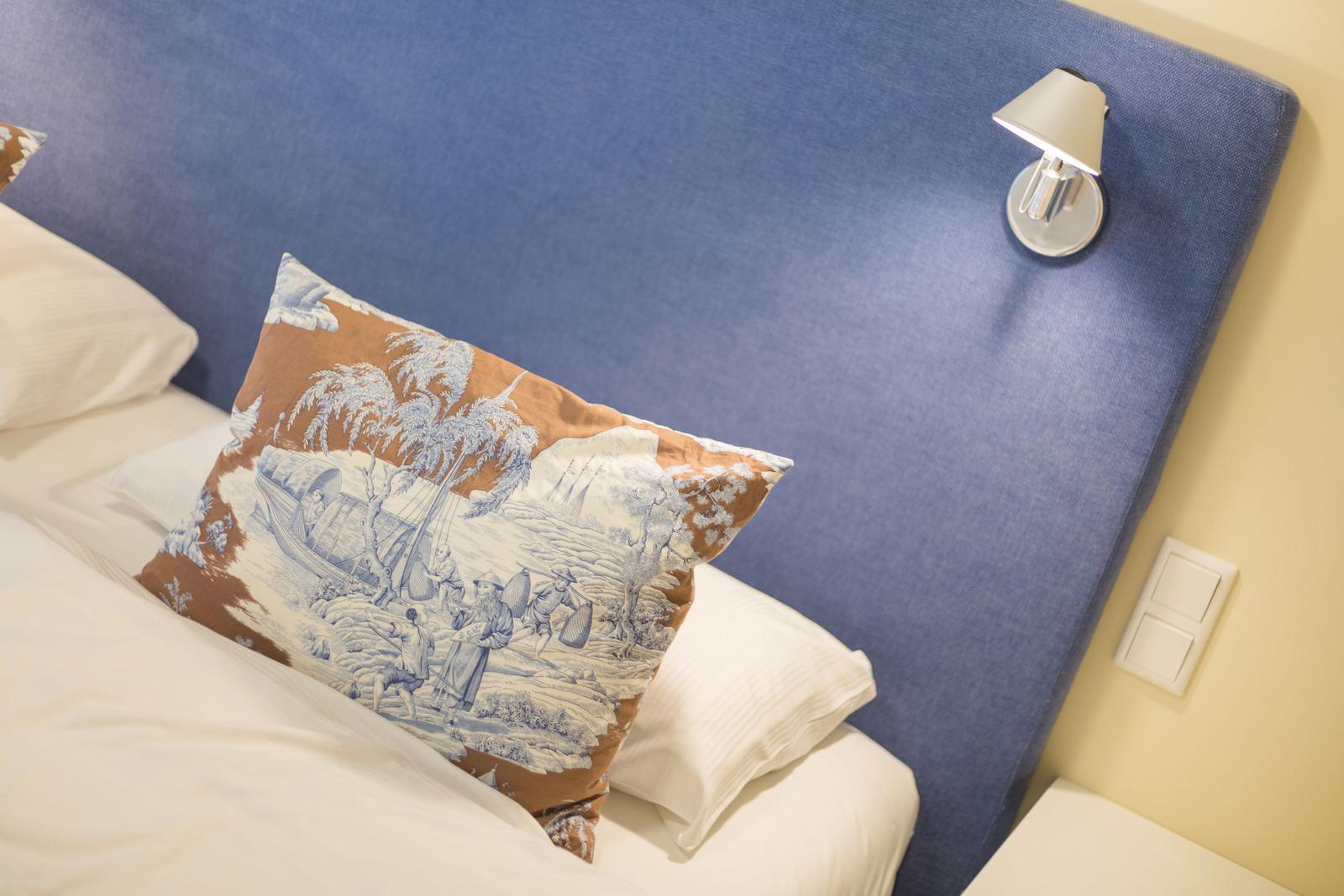 Bettkopfteil mit blauem Stoff, Kissen mit Landschaftsmotiv und Wandlampe.