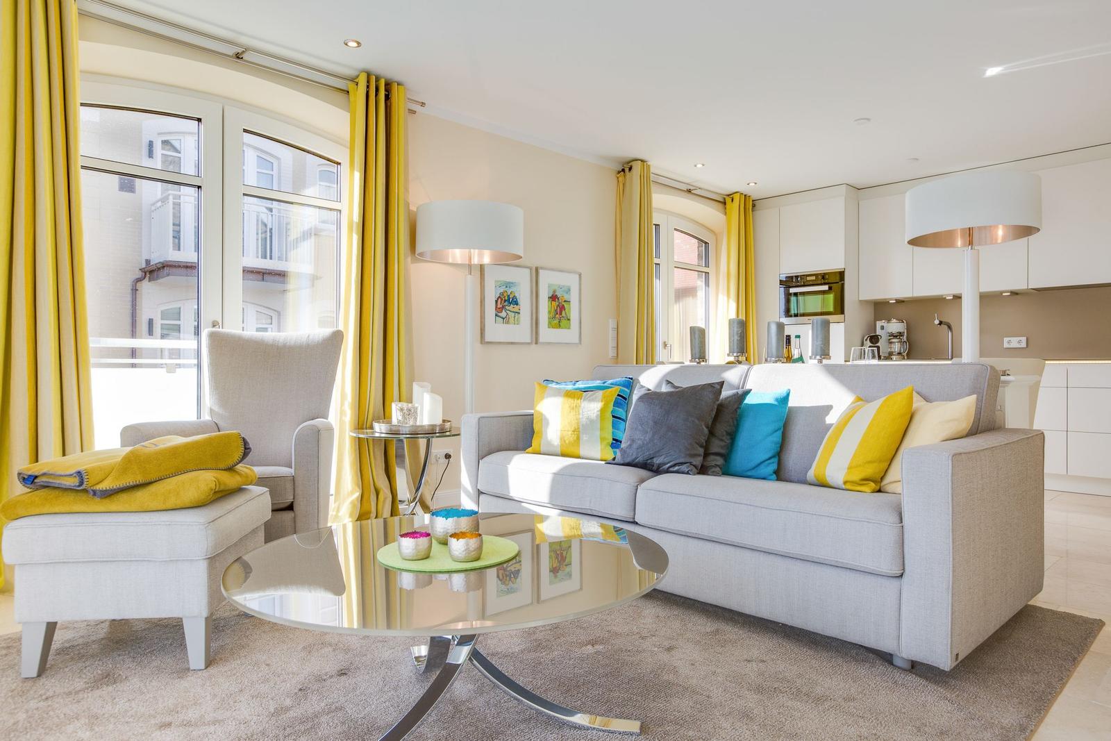 Wohnbereich mit Sofa, Sessel und Glas-Tisch. Gelbe Vorhänge und Küchenbereich sichtbar.