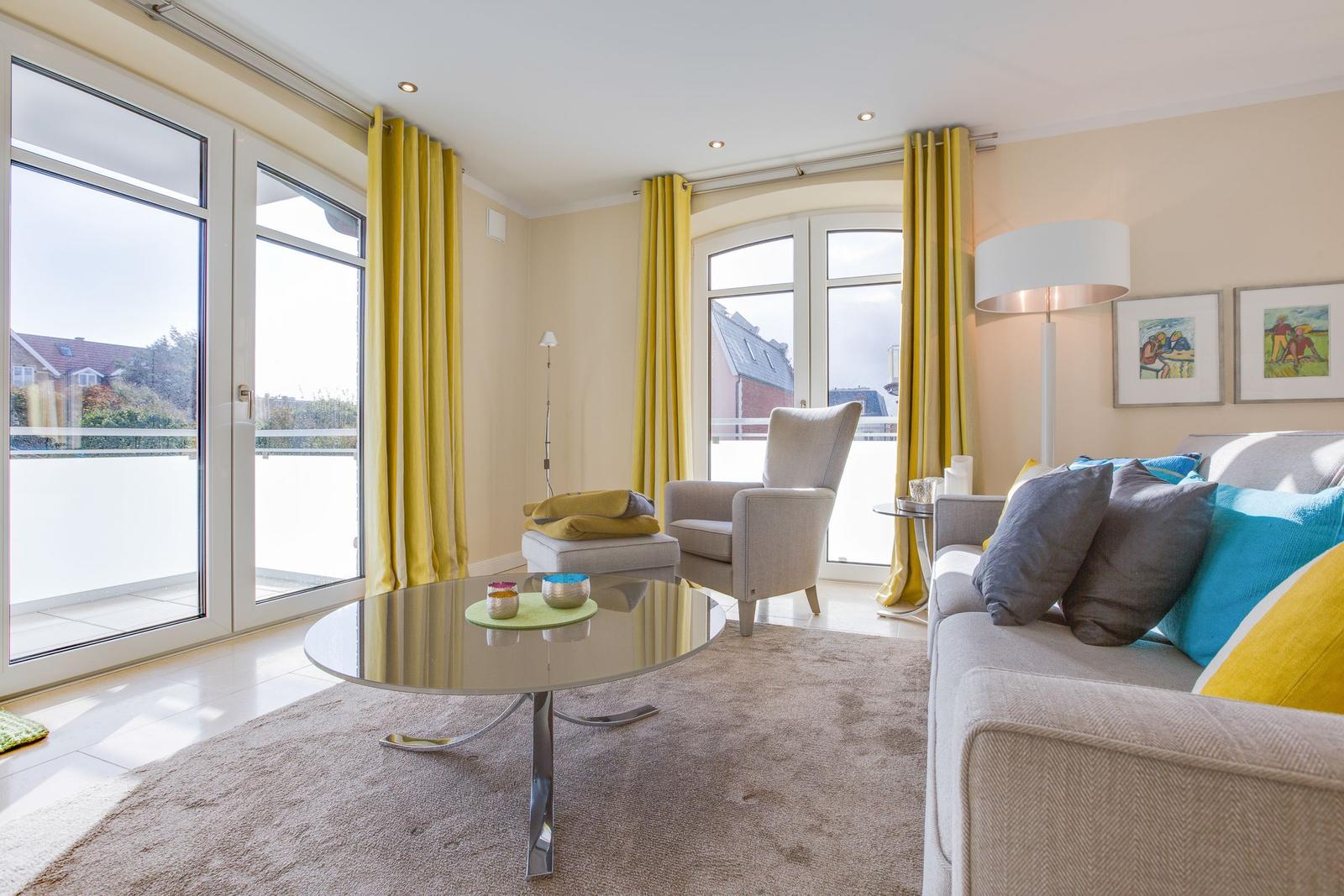 Wohnzimmer mit Sofa, Sessel, Glas-Tisch und gelben Vorhängen.