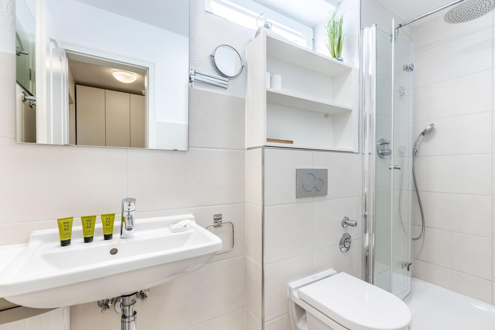 Badezimmer mit Dusche, Waschbecken und Spiegel. Drei grüne Flaschen auf dem Waschbecken.