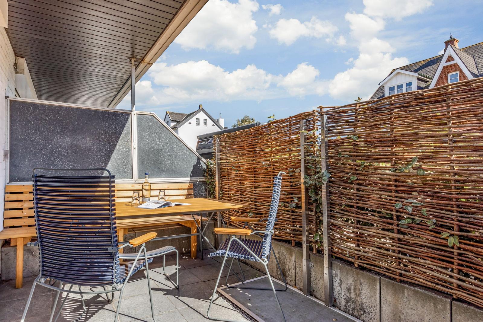 Terrasse mit Tisch, zwei Stühlen und Holz-Zaun. Hintergrund: Häuser und blauer Himmel.