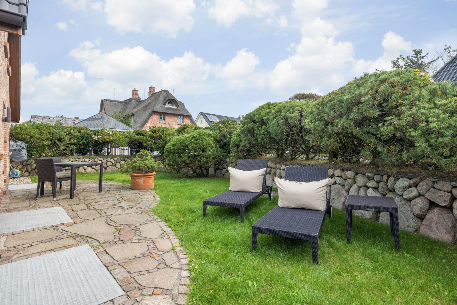 Backyard with loungers, table, and green areas. Stone wall and trees in the background.