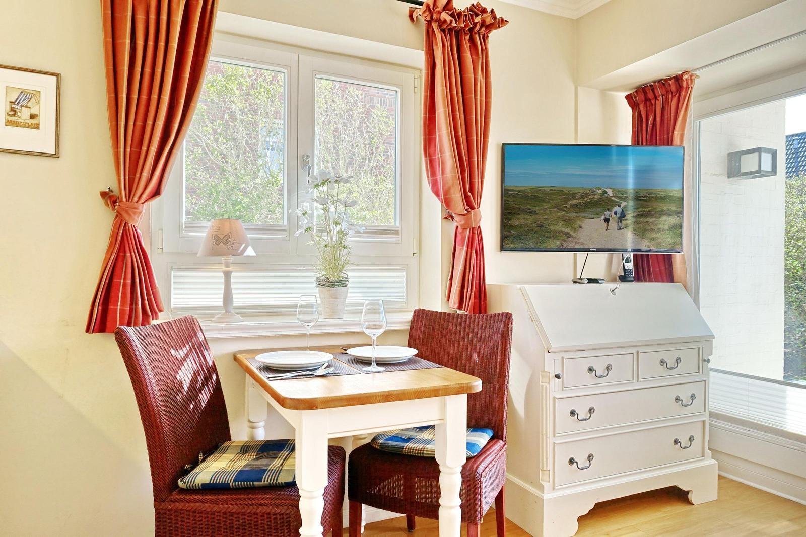Essbereich mit Tisch, Stühlen und TV. Fenster mit roten Vorhängen.