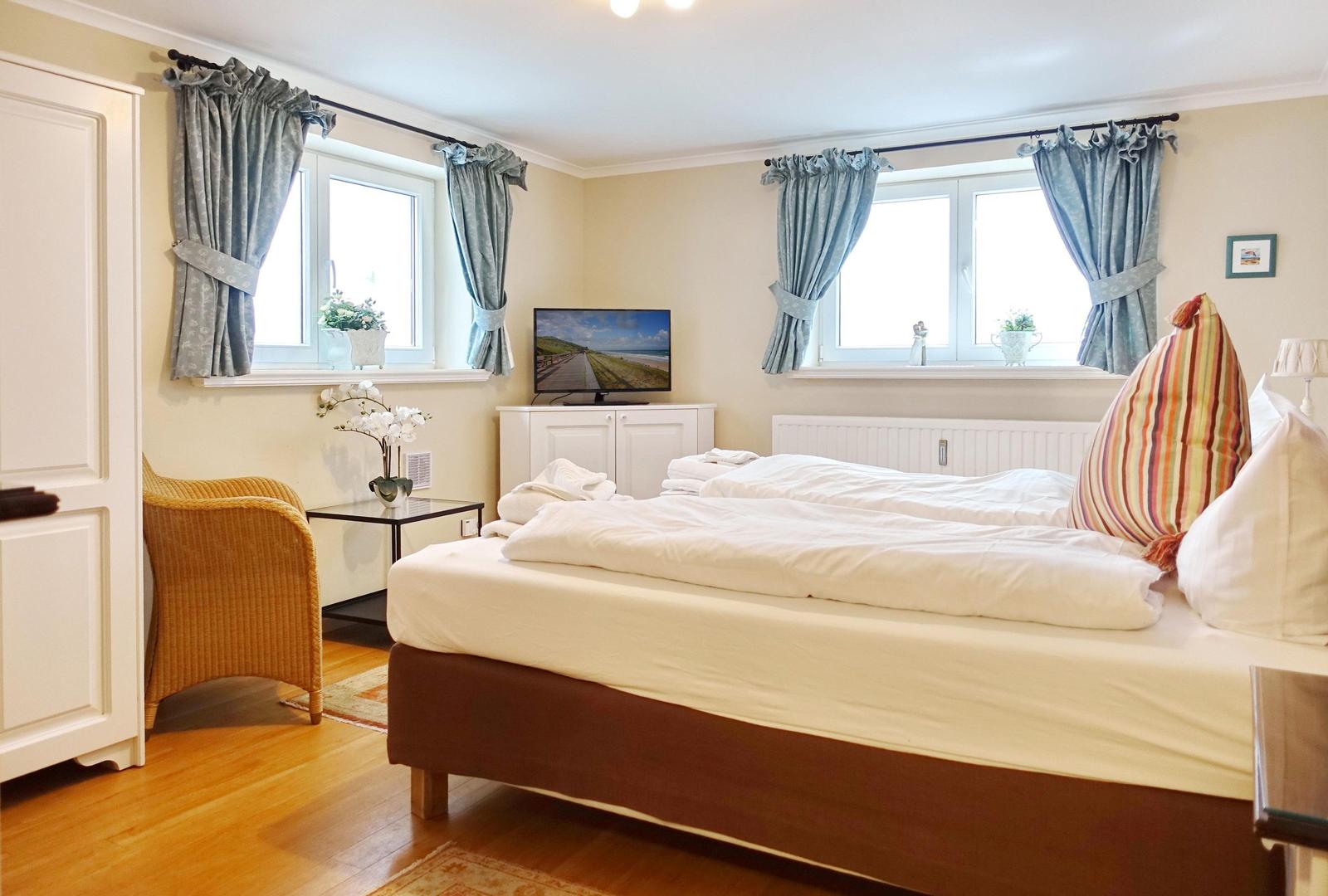 Schlafzimmer mit Doppelbett, zwei Fenstern und Fernseher.
