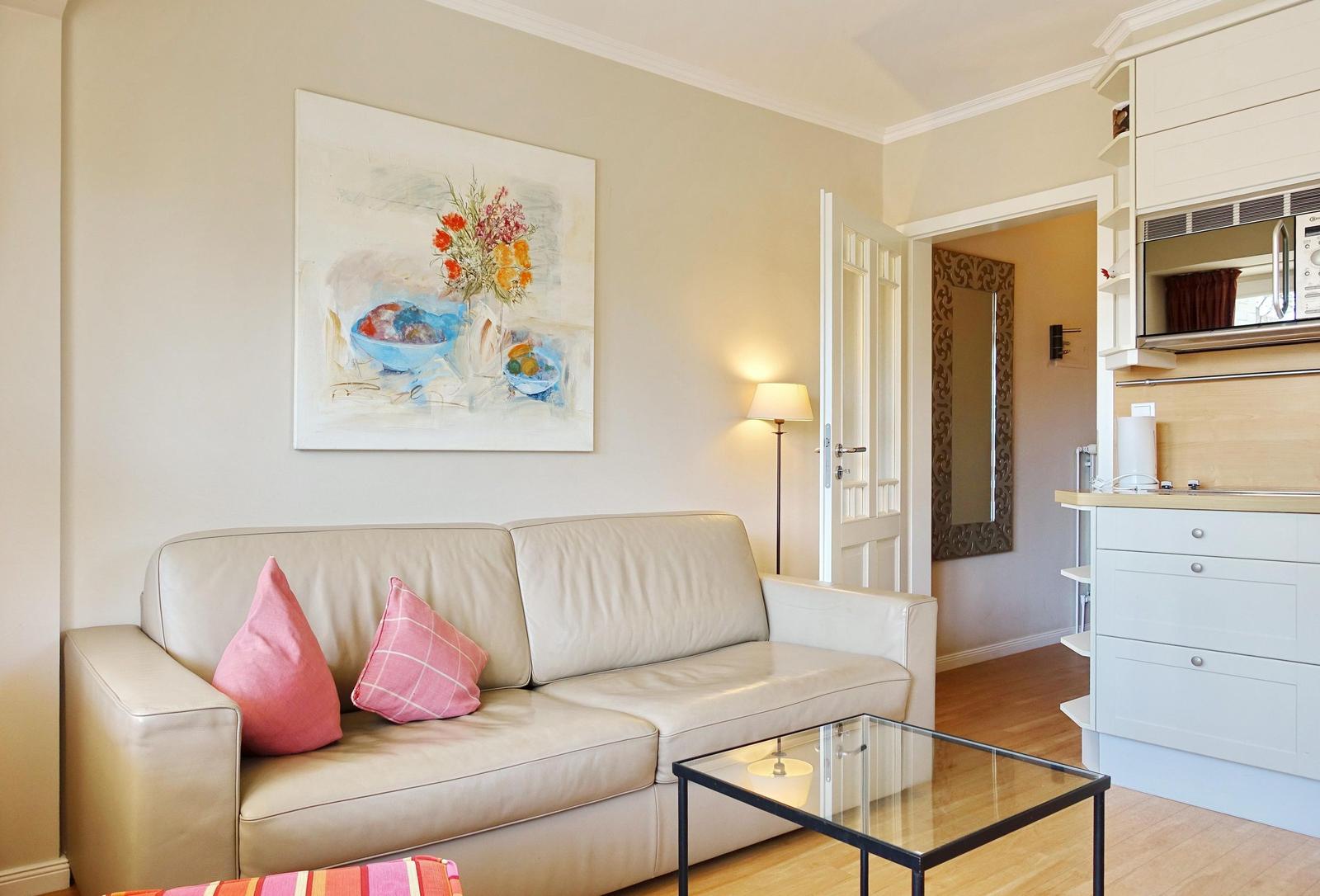 Wohnzimmer mit Sofa, Tisch und Wandbild. Küche mit Mikrowelle sichtbar.