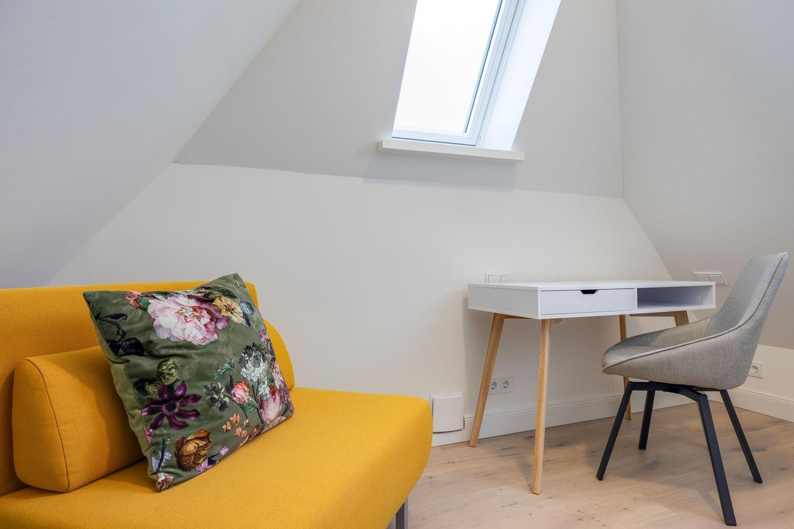 A room with a yellow sofa, a desk, and a window.