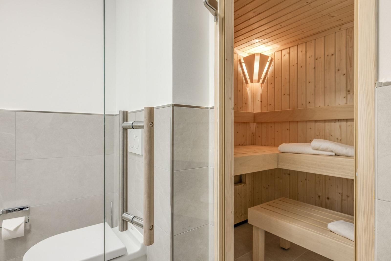 Shower with glass wall and wooden handle. Next to it, a sauna with wooden benches and towel.