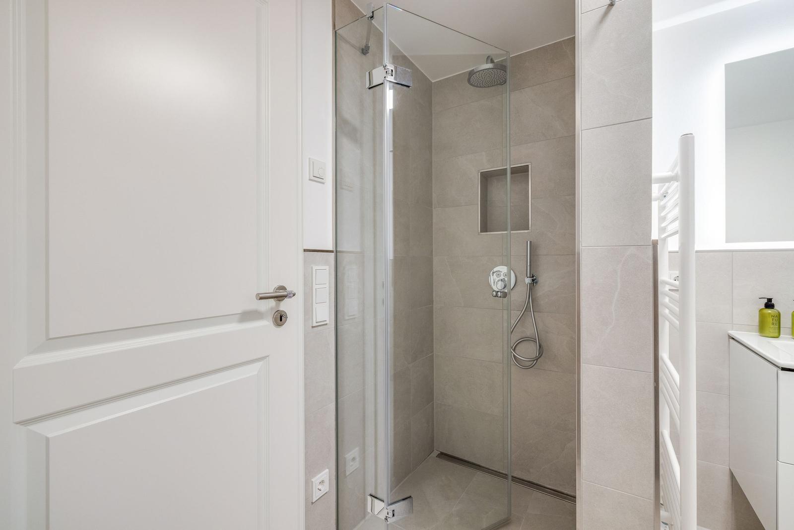 Shower cabin with glass enclosure, showerhead, and handheld shower. White door and vanity with mirror.