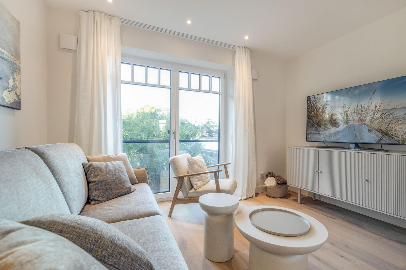 Living room with sofa, armchair, and TV. Large windows with view outside.