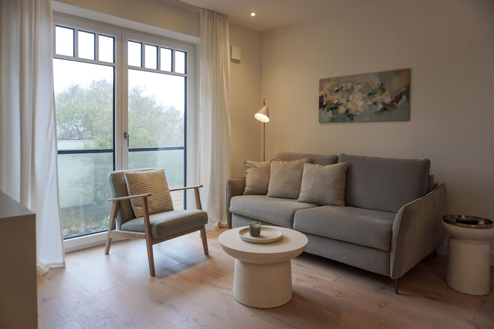 Wohnzimmer mit Sofa, Sessel und Tisch vor großem Fenster.