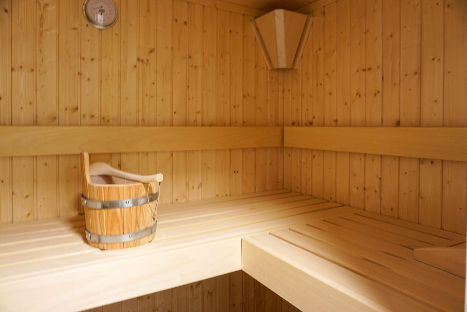 Holzsauna mit Sitzbänken und Eimer. Wandthermometer und Holzverkleidung sichtbar.