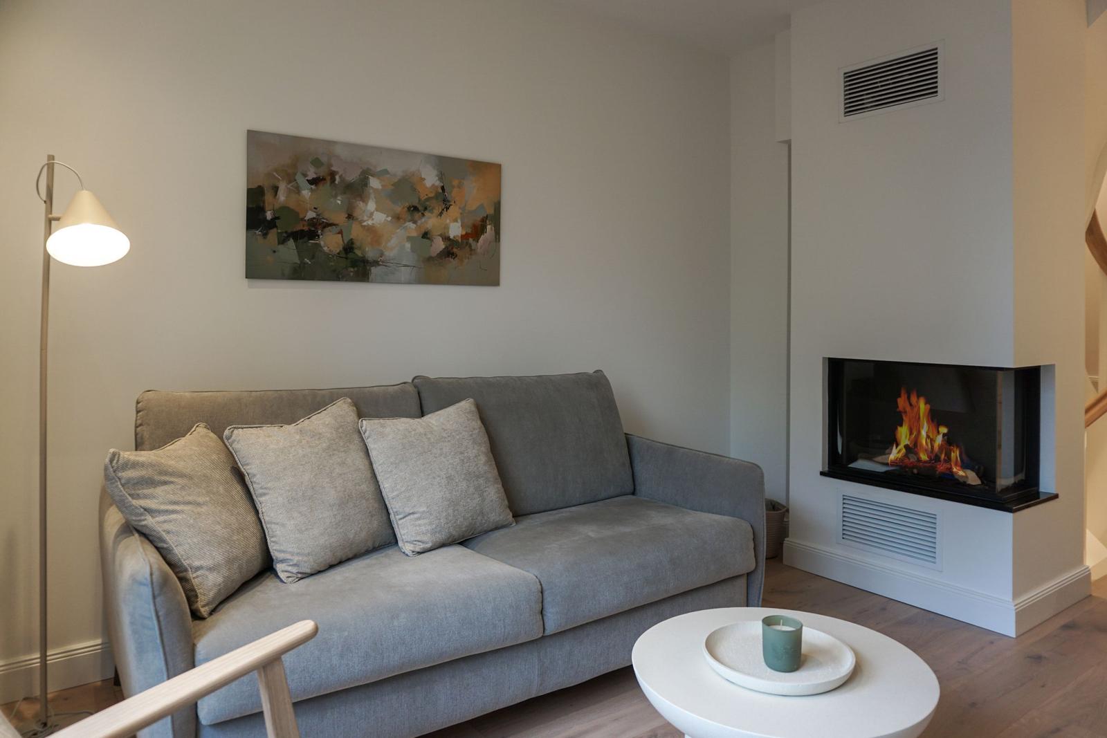 Ein grauer Sofa mit Kissen, eine Stehlampe, ein runder Tisch und ein Kamin mit brennendem Feuer.