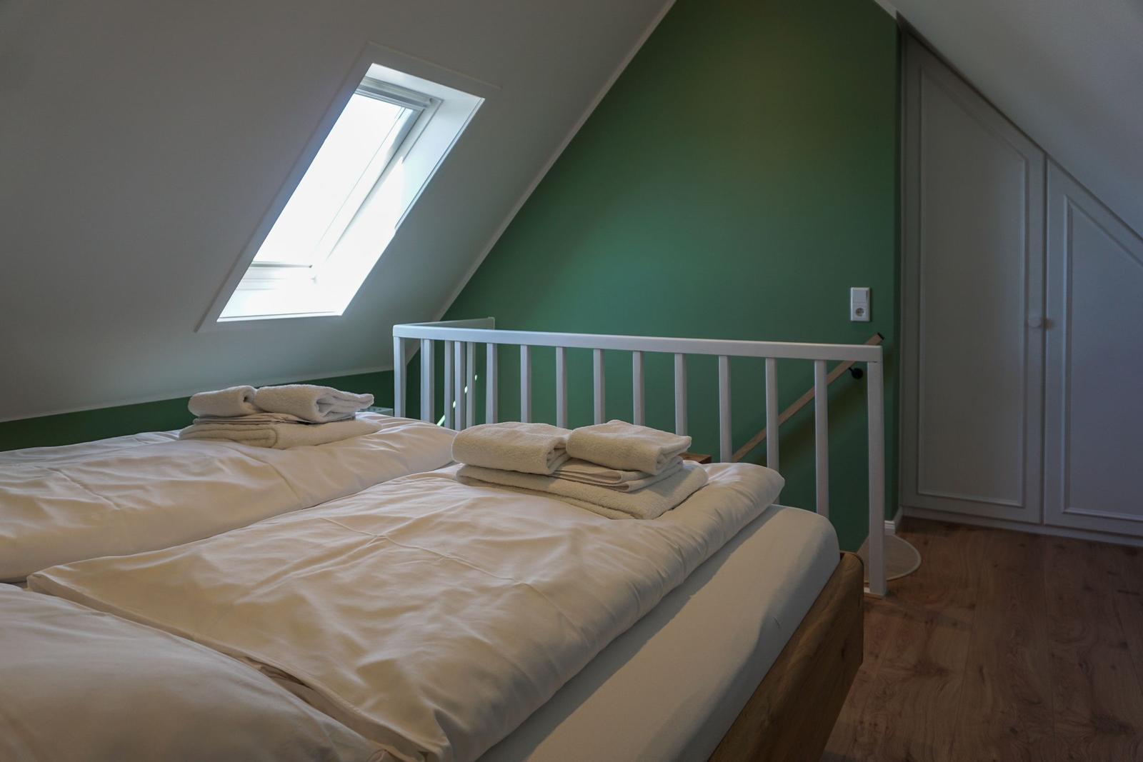 Bedroom with skylight, green wall, and wooden floor. Bed with white sheets and folded towels.