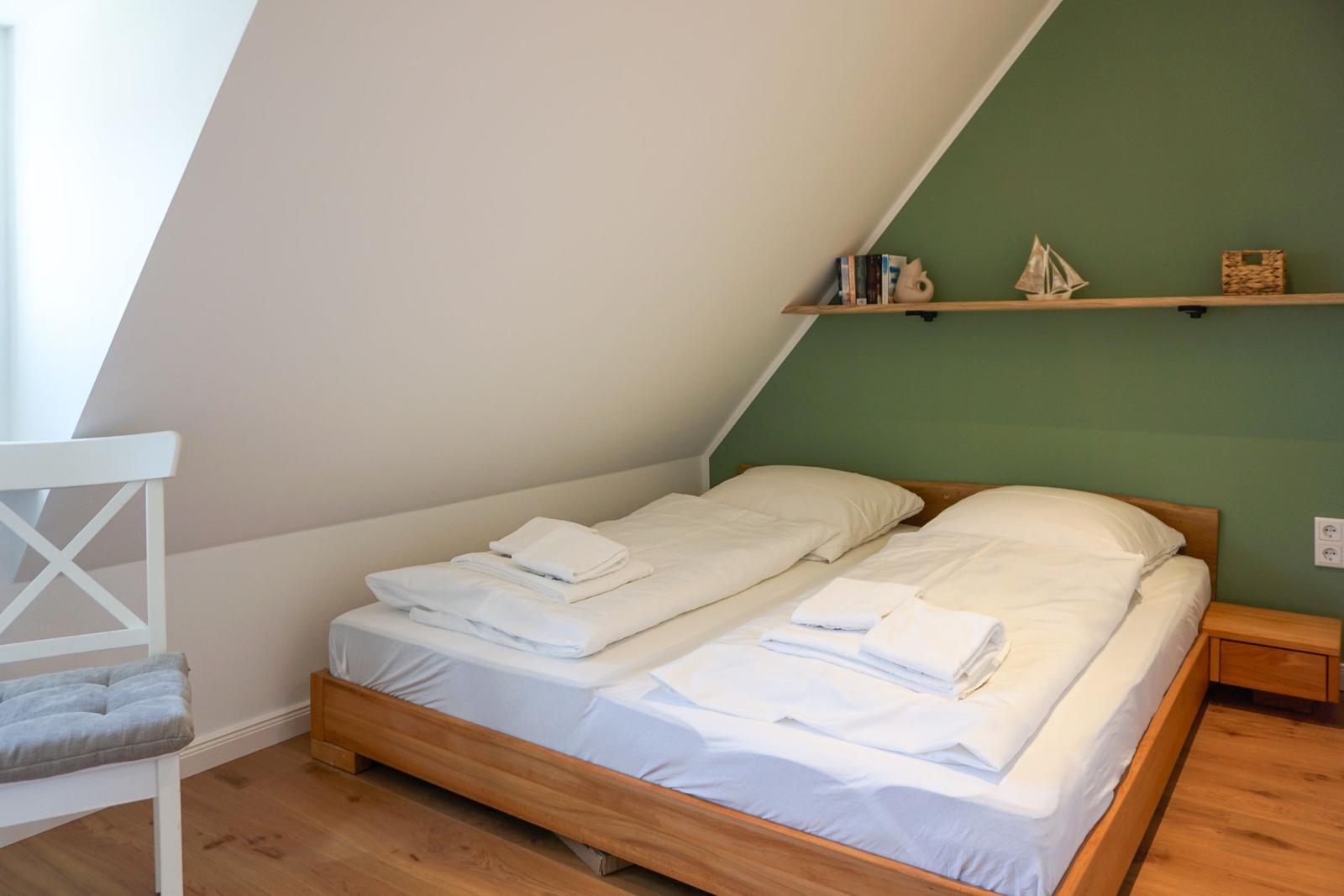 Bedroom with double bed, wooden floor, and green wall. Shelf with decorative items.