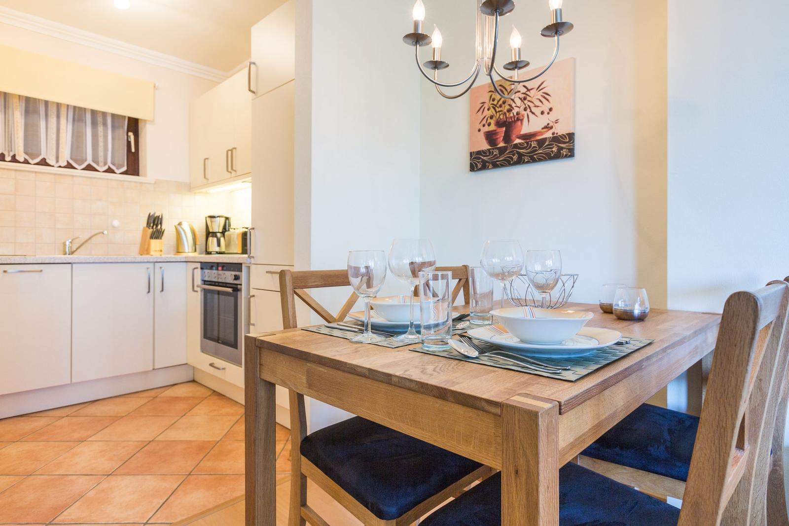 Dining area with wooden table, chairs, and set table. Kitchen in background with countertop and appliances.