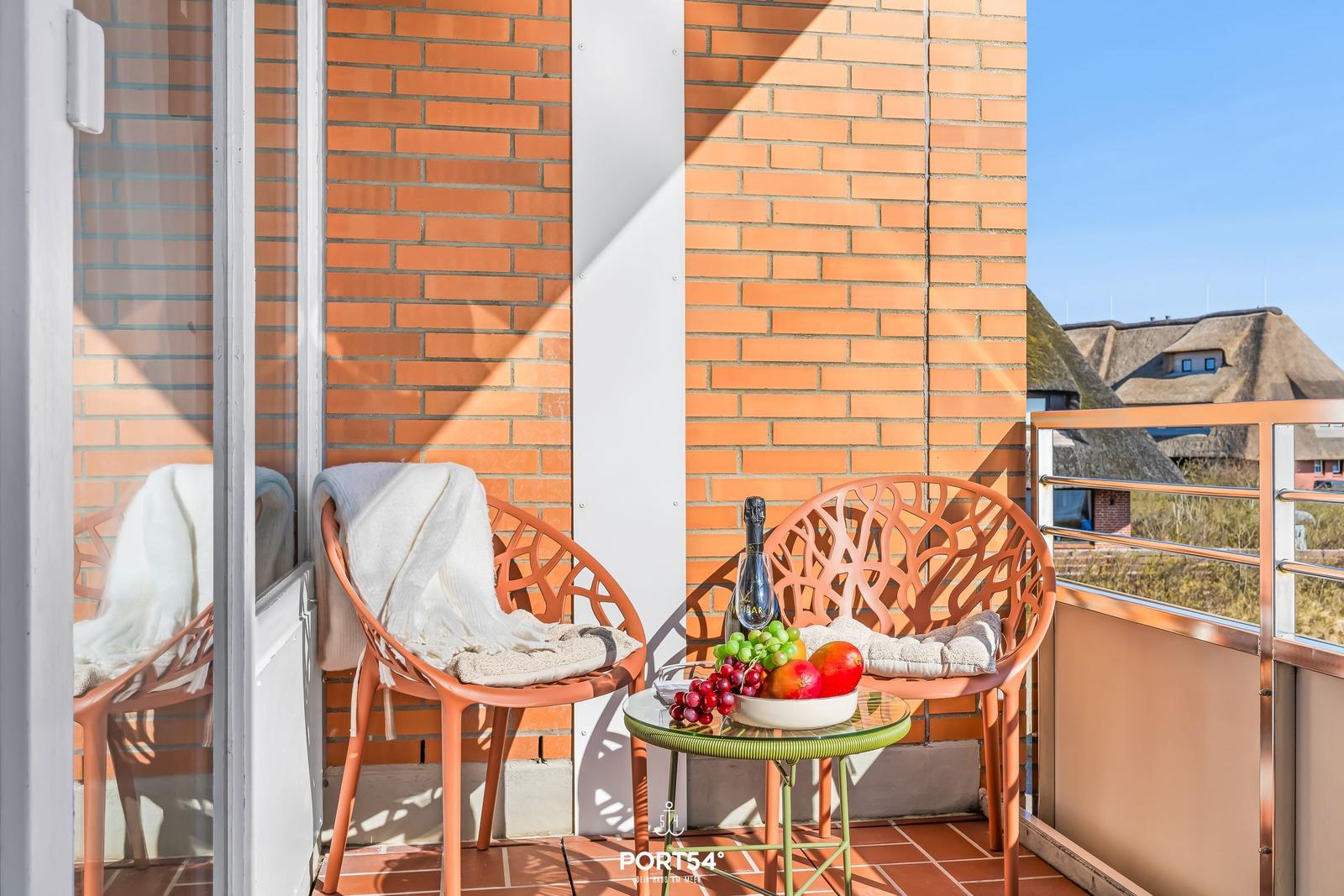 Balcony with two chairs, table, fruit, and bottle. Brick wall and thatched roofs visible.