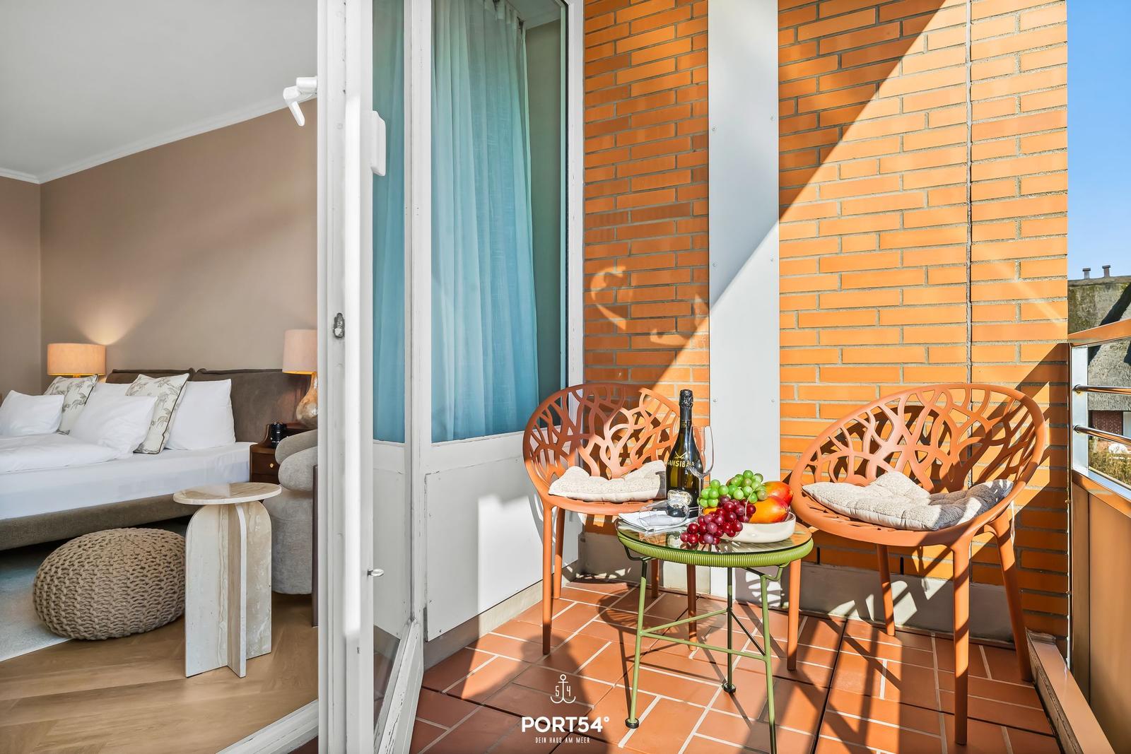 Balcony with two chairs and table, fruit and bottle. Bedroom visible through open door.
