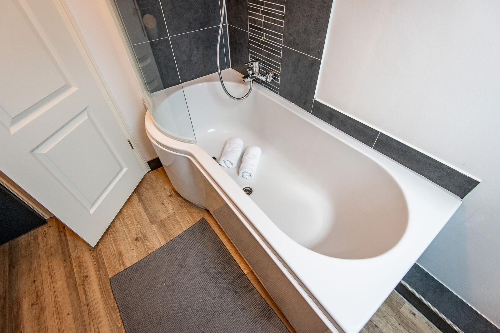 Bathtub with shower screen, two towels, and wooden floor.