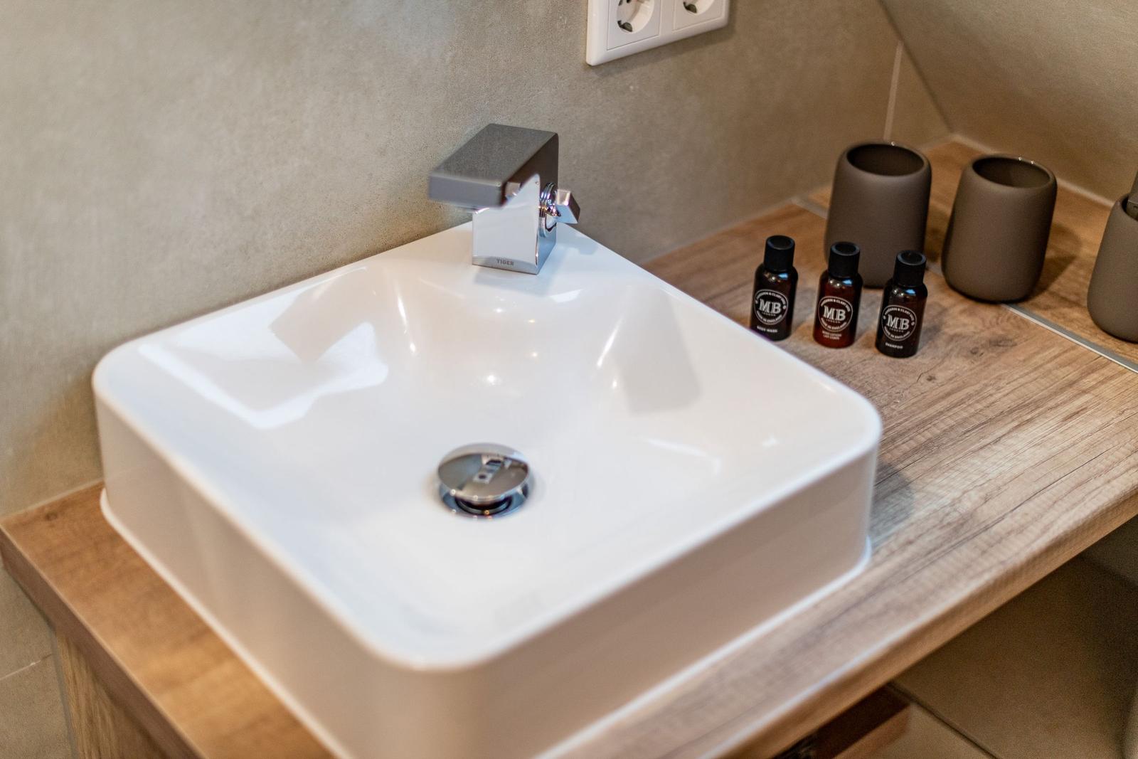 Sink with faucet on wooden vanity, three bottles and two cups.